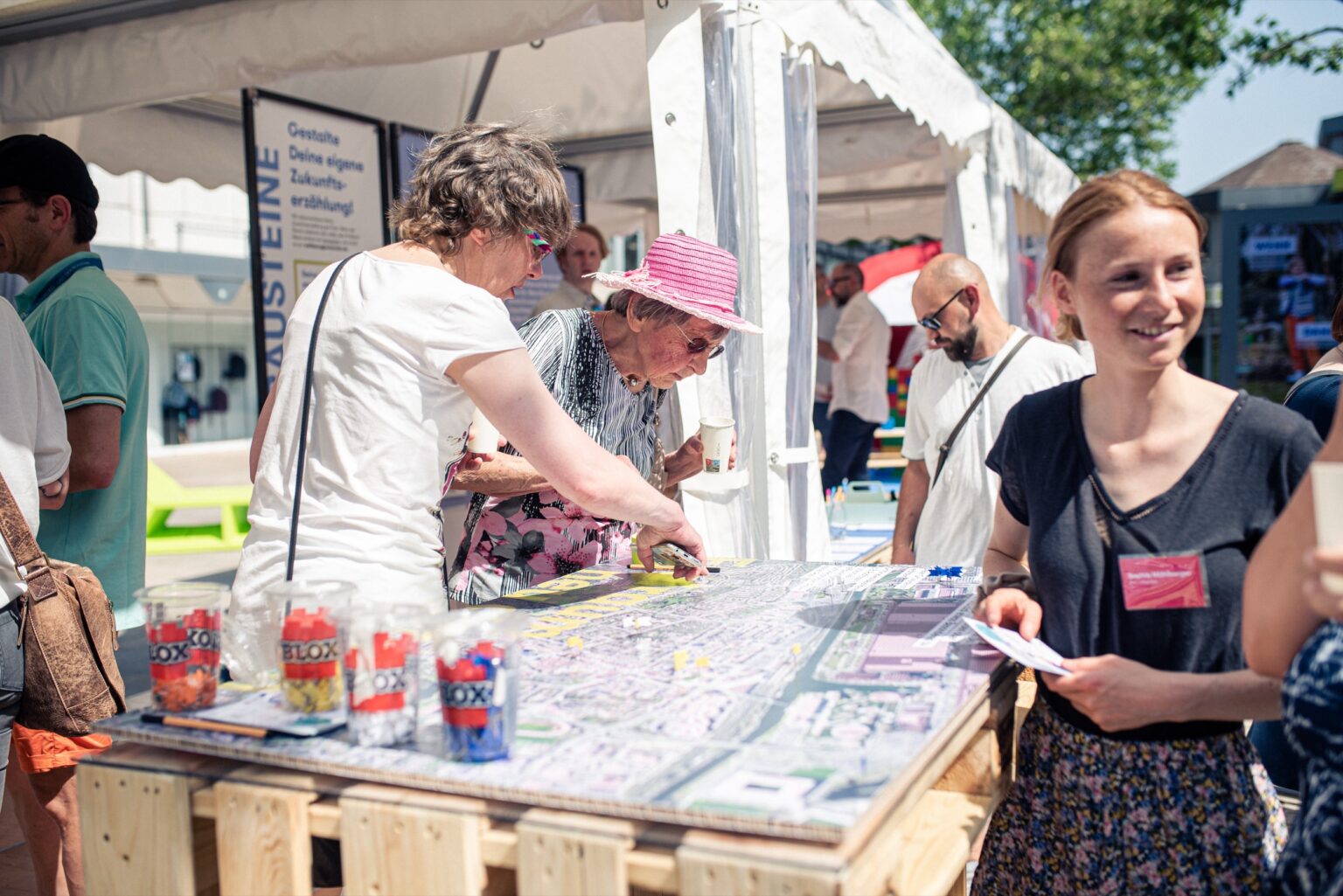 Eine Beteiligungsveranstaltung im öffentlichen Raum an einer Stadtkarte. Zwei Frauen arbeiten an der Karte mit Pins und diskutieren. Im Vordergrund steht eine lächelnde Frau. Im hintergrund sieht man ein großes Veranstaltungszelt mit weiteren Menschen und Stellwänden.