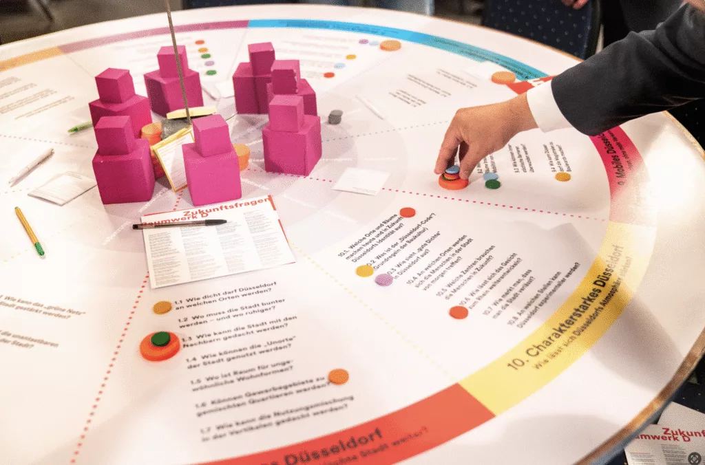 Workshop im Projekt Raumwerk D in Düsseldorf: Teilnehmende legen Spielsteine und Karten auf einen großen runden Tisch mit Zukunftsfragen und pinkfarbenen Modellbausteinen.