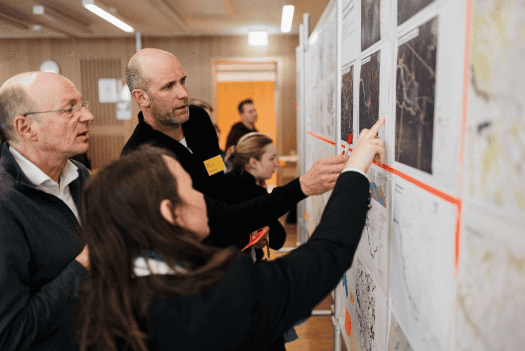 Teilnehmende eines Workshops stehen vor einer Wand mit Karten und Plänen und diskutieren gemeinsam Details, auf die eine Person zeigt.