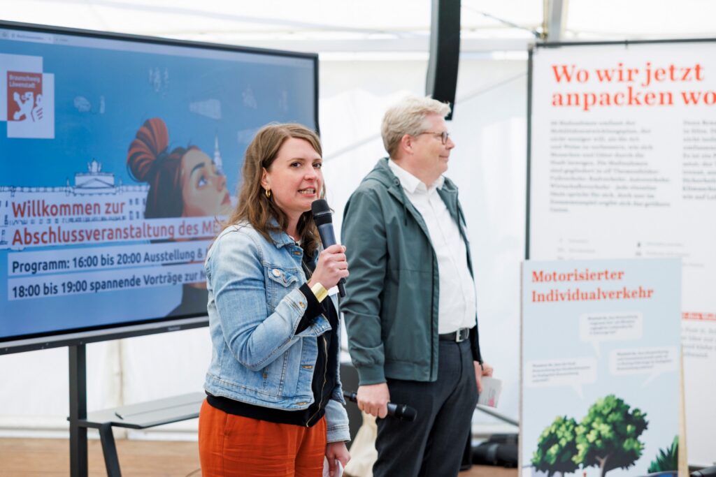 Abschlussveranstaltung des Mobilitätsentwicklungsplans in Braunschweig: Zwei Personen sprechen mit Mikrofonen vor Publikum, im Hintergrund ein Bildschirm mit Programminformationen.