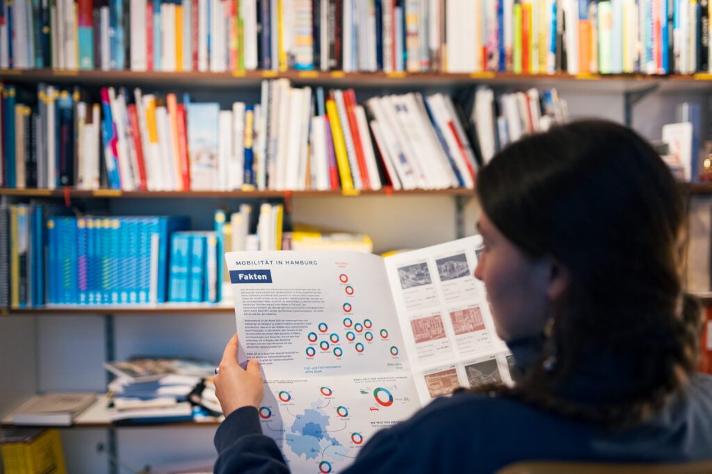 Teammitglied von urbanista sitzt vor einem Bücherregal und liest eine Publikation mit Infografiken zur Mobilität in Hamburg.