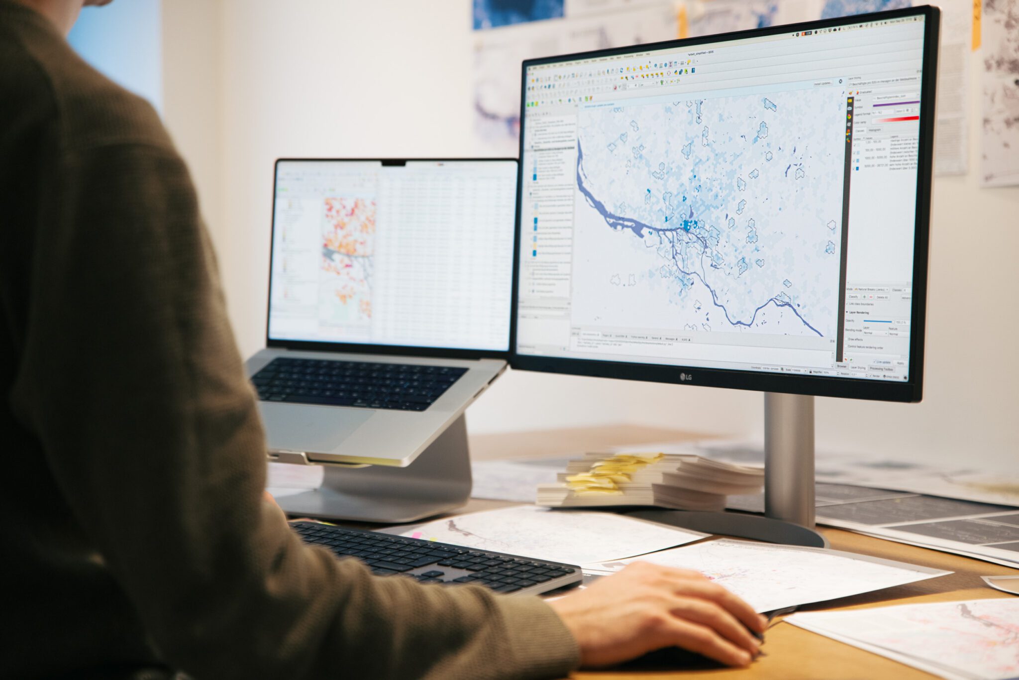 Person arbeitet am Computer mit zwei Monitoren, auf denen Karten und Datenanalysen angezeigt werden.