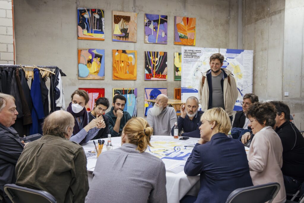 Gruppe von Menschen sitzt um einen Tisch in einem Workshopraum mit bunten Bildern an der Wand und diskutiert über Karten und Pläne.