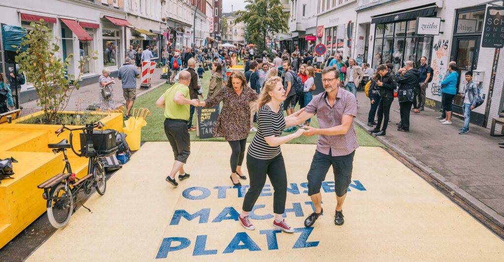 Menschen, die feiern und tanzen bespielen einen Straßenraum neu. Auf der Straße ist ein großer Aufdruck "Ottensen macht Platz". Im Vordergrund tanzen mehrere Paare Standardtanz